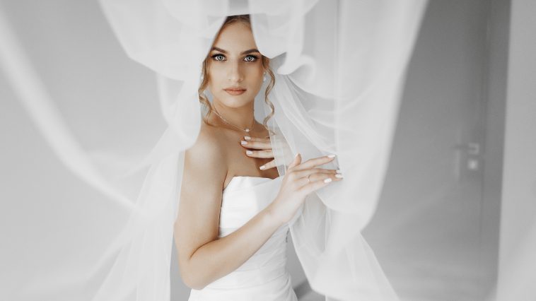 A woman in a white wedding dress is standing in front of a curtain. She is wearing a veil and has her arms crossed. The image has a romantic and intimate mood