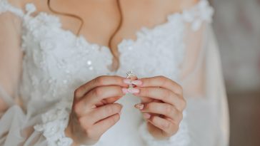 A woman is holding a ring in her hand. The ring is silver and has a diamond on it. The woman is wearing a white dress, and the dress is long and flowing. Concept of elegance and sophistication