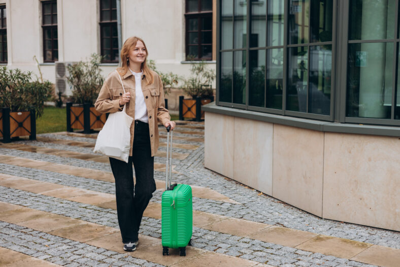 Cheerful young woman in casual outfit walking on the city street with green luggage. 30s Woman dragging suitcase on urban background. Travel Fashion.