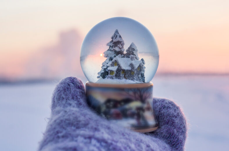 Girl in knitted mittens holding glass ball with firtrees, house and artificial snow with winter landscape at the background