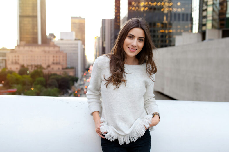 Beautiful young woman sitting on a bridge across the boulevard in urban scenery, downtown, at sunset, smiling at camera.