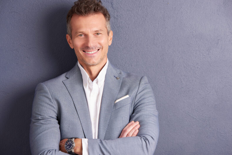 Close-up portrait shot of confident middle aged businessman with folded arms standing at grey wall and looking at camera.