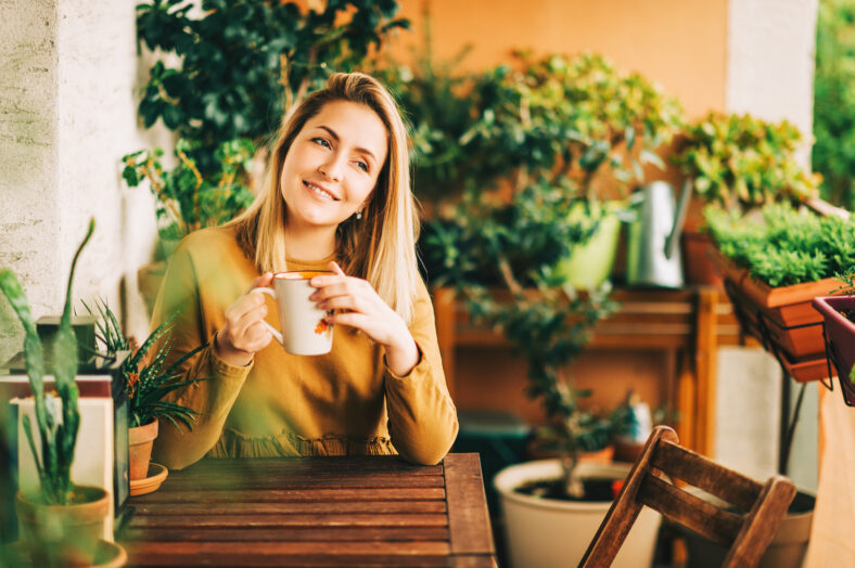 Romantic portrait of beautiful young woman wearing brown cotton dress, relaxing on the balcony between many green plants, holding cup of tea or coffee