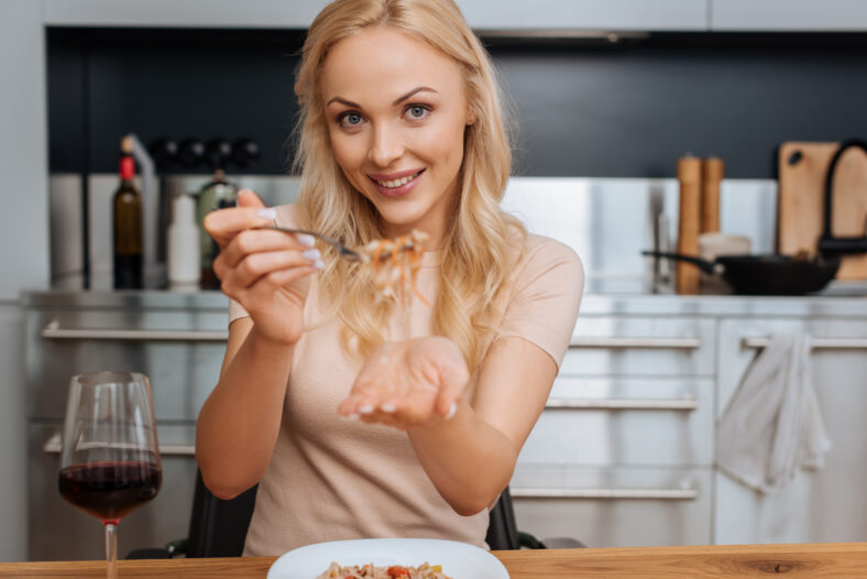 cheerful woman holding fork with thai noodles while smiling at camera near glass of red wine