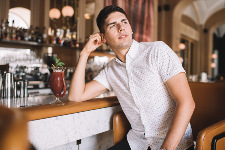 Pensive young brown haired man in casual wear enjoying red cocktail and looking away while spending time in restaurant with bar counter