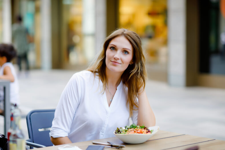 Young beautiful woman eating fresh salad in outdoor restaurant. Woman enjoying lunch bowl with fresh vegetables. Healthy food. On summer day.