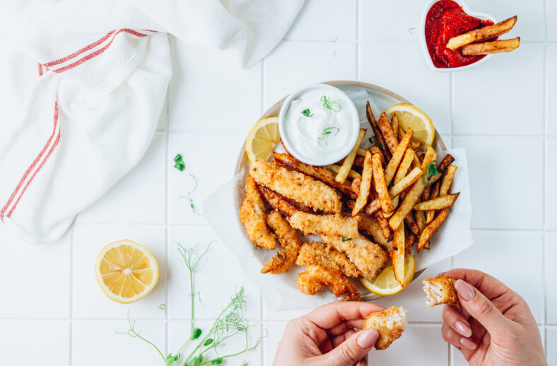 British Traditional Fish and chips with ketchup and tartar sauce on white plate. Woman's hand holding fish. White tiled background. Top view