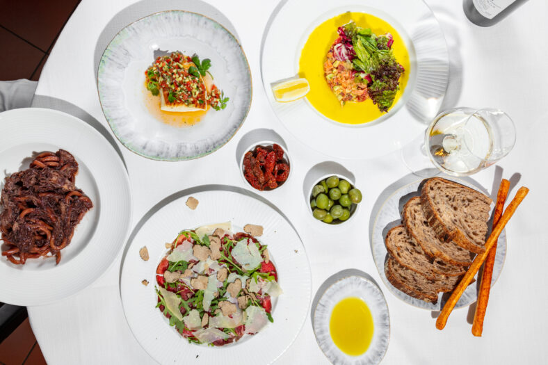 Lunch of Italian cuisine. There are four plates of dishes on a table with a white tablecloth. Bucatini pasta with duck fillet in plum sauce, salmon tartare, perch fillet with tabouleh and veal carpaccio with truffle. The food lies in light, round ceramic plates. Next to the plates is a bowl of dried tomatoes, a bowl of olives, a plate of buckwheat bread and gressini, a saucer of olive oil and a glass of white wine. Top view.