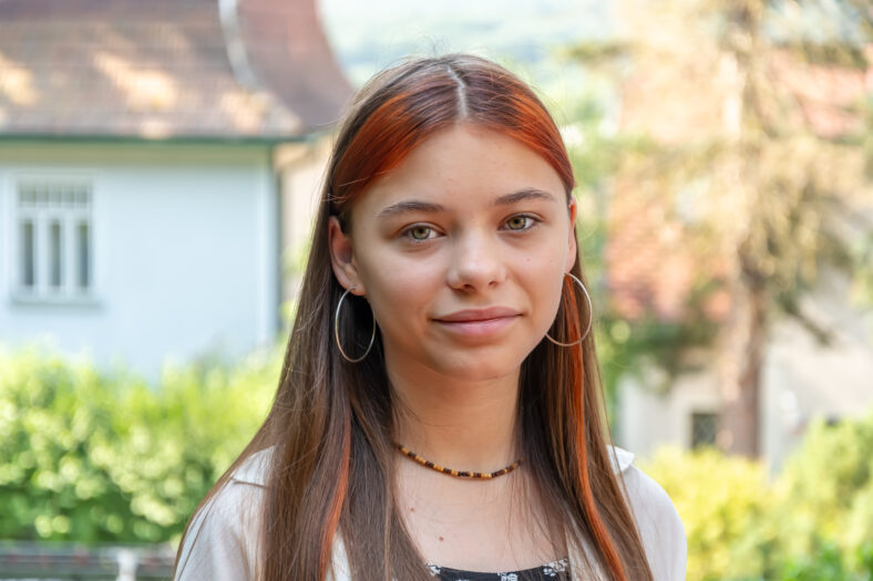 A young woman with long brown hair with red strands and hoop earrings looks directly into the camera. The background is blurred, with the greenery, house and tree trunk in focus.