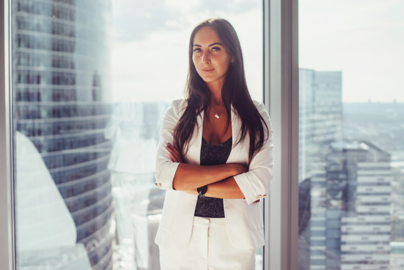 Portrait of elegant business lady wearing white formal suit standing near window looking at camera.