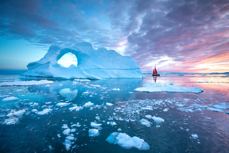 Little red sailboat cruising among floating icebergs in Disko Bay glacier during midnight sun season of polar summer. Ilulissat, Greenland.