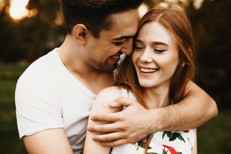 Portrait of a amazing couple laughing while man is embracing from back her girlfriend with red hair against sunset while dating in their vacation time.