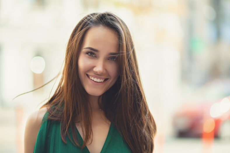 Young pretty likable cheerful woman posing summer city outdoor. Beautiful self-confident girl dressed in emerald-colored jumpsuit with long brown hair walking street enjoing her life, urban lifestyle