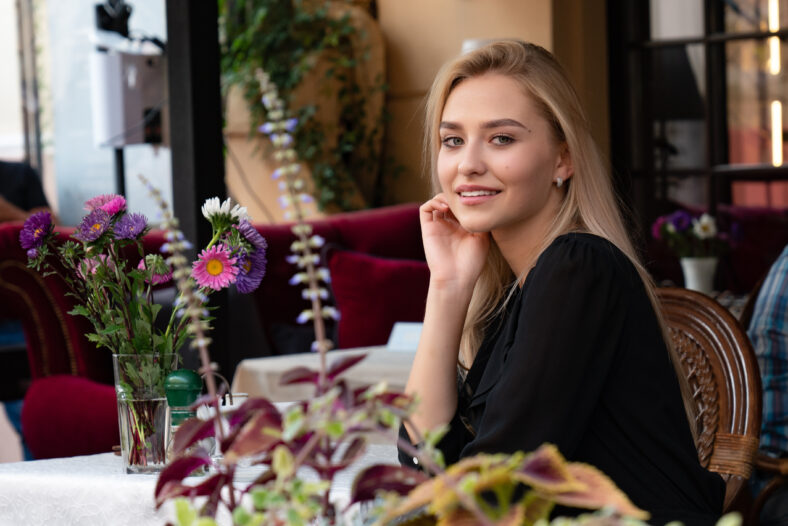 Elegant lady with blonde loose hair and natural makeup, dressed in black dress sitting at a table in a cozy street cafe decorated with flowers, outdoor photo