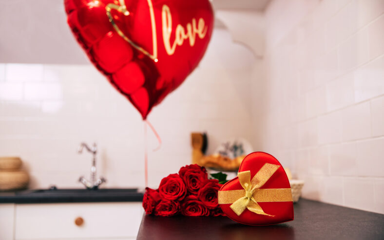 Close-up photo of a bouquet of red roses, a big balloon and a red heart-shaped box with a golden ribbon, which lie on a kitchen table.