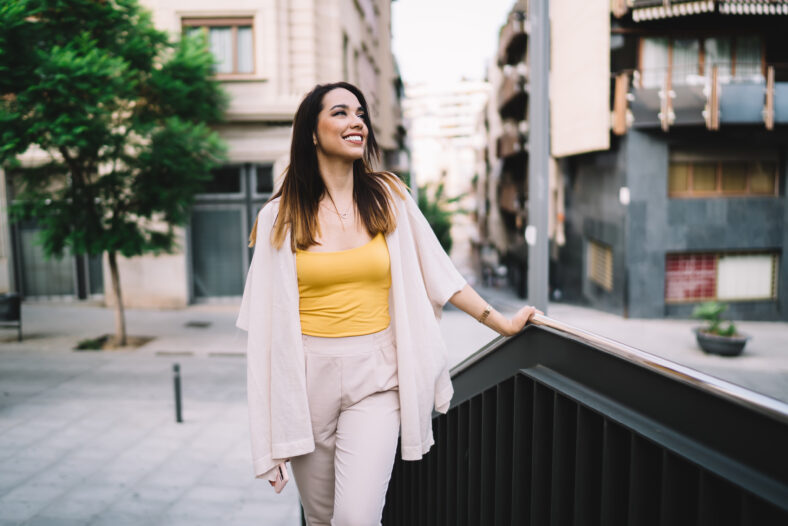 Toothy hipster girl dressed in stylish outfit laughing at urban street during travelling time, happy Caucasian female tourist in trendy clothing feeling good during solo vacations for recreating