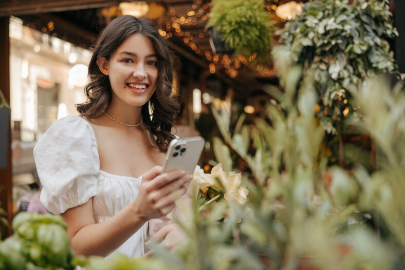 Smiling young caucasian woman looking at camera, holding phone standing outdoors. Brunette spends her leisure time among plant market. Concept of enjoying moment, technology