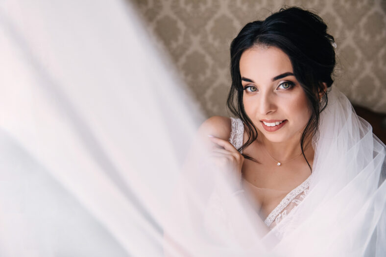 A beautiful, sweet, bride in a white lace dress with a veil is standing by the window against the wall. Wedding model portrait.