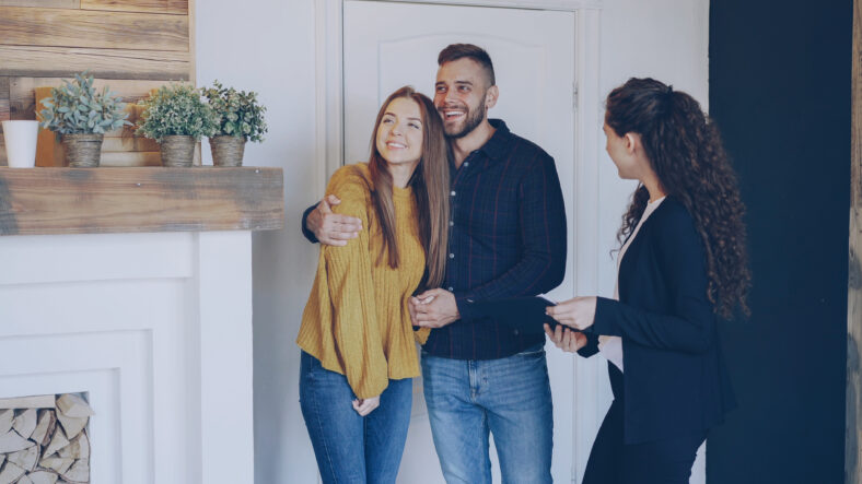 Real estate agent is meeting happy married couple, showing papers and telling them about new apartment. Young people are excited, they are looking around and hugging.