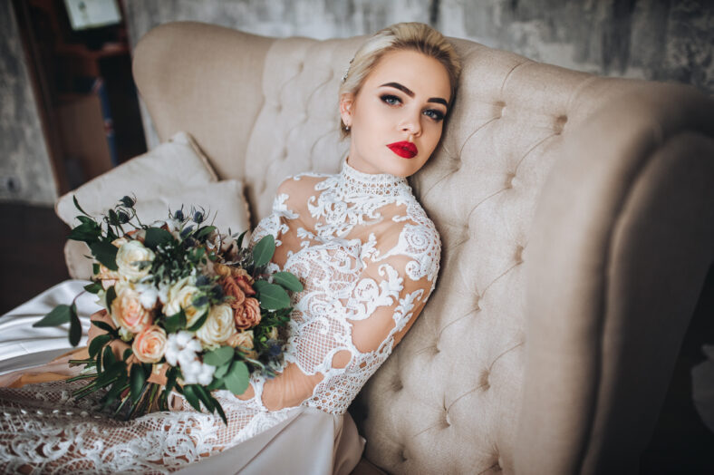 Beautiful bride in the original lace wedding dress sits on the couch with a bouquet in hands.