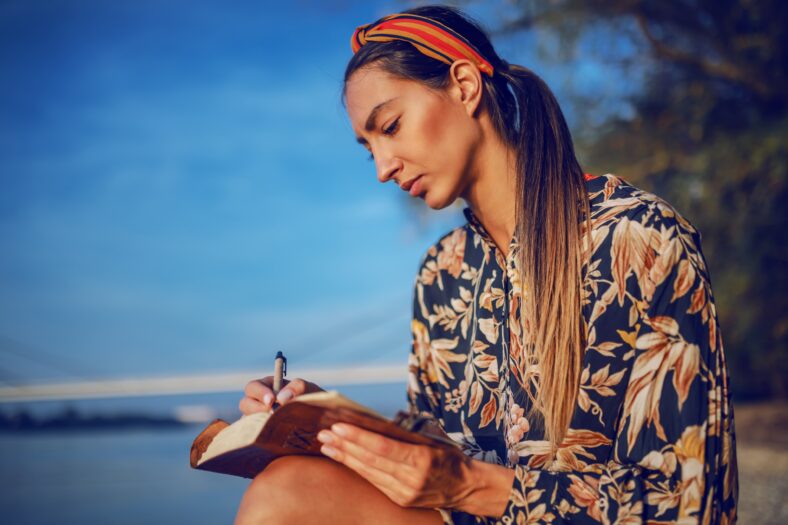 Portrait of pensive attractive caucasian brunette in floral dress and with headband sitting on rock on shore next to river and writing diary.