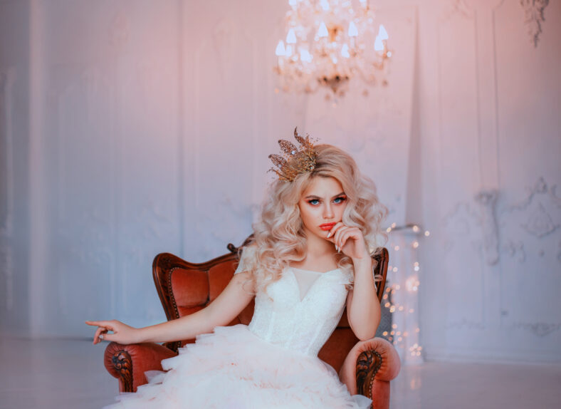 Angry young beautiful girl princess misses sadness loneliness. Queen woman blonde woman long curls hair. Hairstyle with vintage royal crown. Backdrop white room, candelabra romantic lit, bright sparks