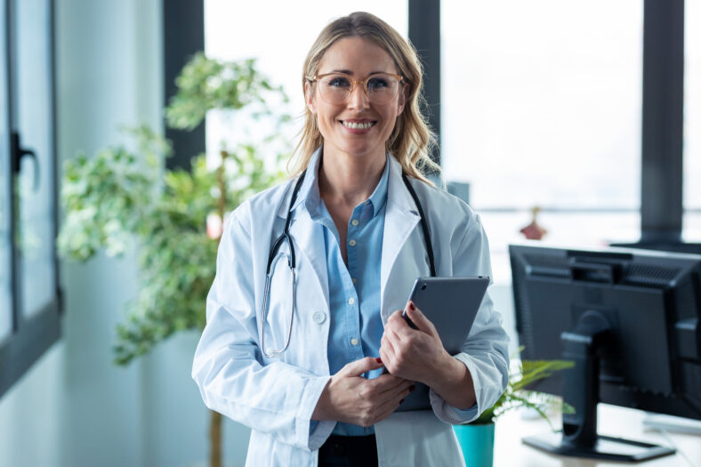 Retrato de atractiva doctora madura que mira la cámara mientras sostiene la tableta digital en la consulta.