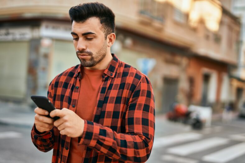 El joven hispano con una expresión seria usando teléfonos inteligentes en la ciudad.