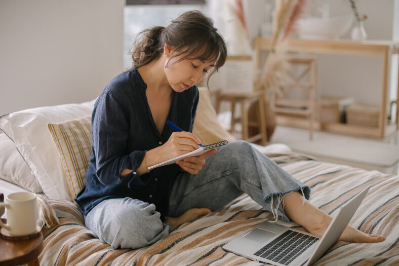 Asian young woman making notes in notebook, working at home on bed in cozy bedroom. Concept of freelancer, remote work, home office.