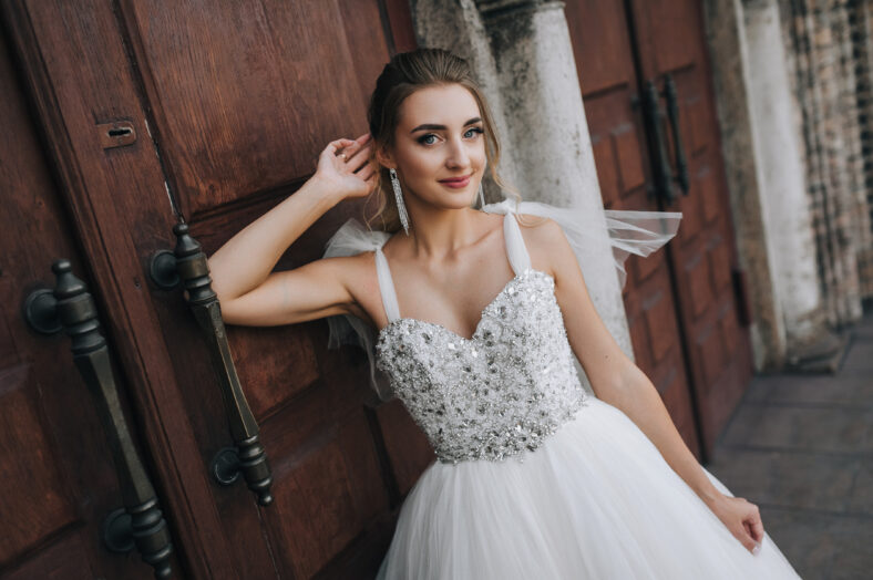 Beautiful smiling bride curly blonde in white lace long dress stands, walks against the background of an old retro church, wooden doors. Wedding photography, portrait.