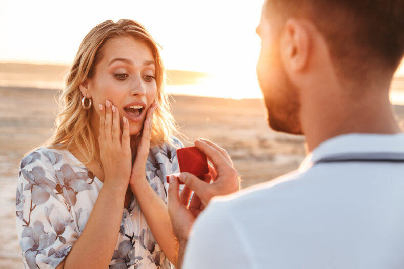 Photo of unshaven man making proposal to his amazed woman with ring in gift box while walking on sunny beach